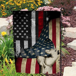May include: A garden flag featuring a distressed American flag design with a clenched fist overlay. The flag displays the stars and stripes in red, white, and blue, with a vintage, worn appearance. The fist is rendered in a sketched style.