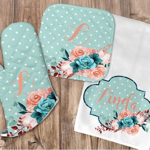 Puede incluir: Conjunto de tres manoplas de horno de lunares azul verdoso y blanco y un paño de cocina con diseños florales y el nombre "Linda" en color naranja melocotón.