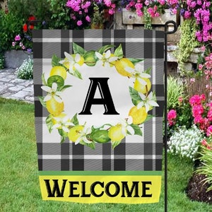 May include: A black and white checkered welcome flag with a wreath of lemons and green leaves. The wreath surrounds the letter "A" in white. The text "WELCOME" is printed in yellow on a green banner at the bottom of the flag.