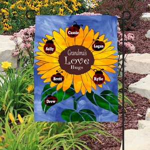 May include: A garden flag featuring a sunflower design with ladybugs and the text "Grandma's Love Bugs." The sunflower has yellow petals and a brown center. Each ladybug has a name on it. The background is blue.