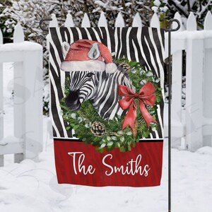 Puede incluir: Una bandera de jardín con temática navideña con una cebra que lleva un gorro de Papá Noel y un lazo rojo. La cebra está rodeada de una corona de vegetación y bayas rojas. La bandera tiene un fondo rojo con el texto "The Smiths" en blanco.
