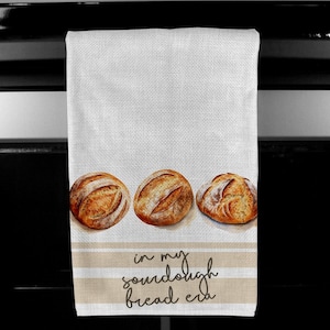 May include: A white kitchen towel with brown stripes and three brown loaves of bread. The text "in my sourdough bread era" is written in black on the towel.