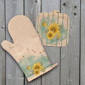 May include: A set of two beige oven mitts with a bee and sunflower design. The mitts have a light blue and green watercolor background with gold glitter accents.
