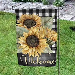 May include: A welcome garden flag with a black and white checkered background. The flag features a watercolor painting of sunflowers with green leaves. The text "Welcome" is written in gold on a black wooden background.