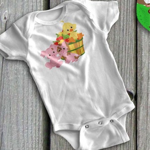 Puede incluir: Body de bebé blanco con una colorida ilustración de un patito amarillo y dos cerdos rosas con manchas marrones. Los animales están cerca de una cesta de madera llena de manzanas y zanahorias. El body tiene mangas cortas y cierres a presión.