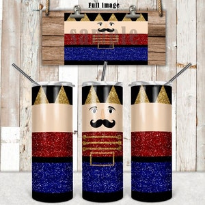 May include: Three stainless steel tumblers with a glitter finish. The tumblers feature a Nutcracker design with a red, blue, and gold color scheme. The design includes a face with a mustache and a gold crown.