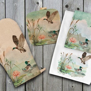 May include: A set of kitchen towels and oven mitts featuring a watercolor design of ducks and cattails. The design is in shades of green, brown, and pink.