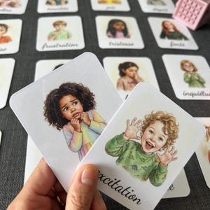 Op de afbeelding: Emotie flashcards met illustraties van kinderen. De kaarten tonen emoties als "opwinding" en "frustratie". Een kleine roze kubus is zichtbaar op de achtergrond. De kaarten worden in een hand gehouden.