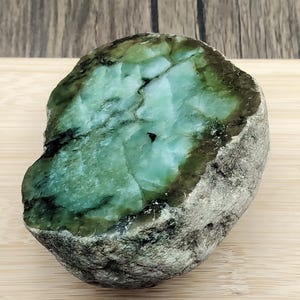 Puede incluir: Una piedra en bruto parcialmente pulida que presenta un interior translúcido de color verde menta vibrante y verde bosque profundo con fracturas cristalinas visibles. El exterior está recubierto por una corteza rocosa natural rugosa, texturizada, de color gris claro y bronceado.