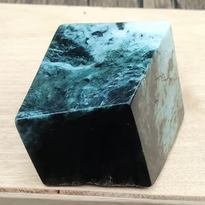 Puede incluir: Un cubo de piedra pulida que presenta un patrón jaspeado en verde bosque profundo y negro con vetas blancas. El objeto tiene bordes afilados y limpios, y una superficie lisa y reflectante que resalta las variaciones de color naturales.