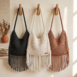 May include: Three macrame shoulder bags in black, cream, and brown hang on wooden pegs. Each bag features a woven body, long fringe, and a braided strap. The bags are a bohemian style.