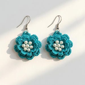 May include: A pair of dangle earrings featuring crocheted teal blue flowers with layered petals. Each flower center is embellished with a cluster of six small, round, white faux pearls. The earrings hang from silver-toned metal fishhook wires.