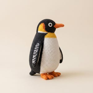 May include: A handmade crochet penguin toy. The penguin has a black head and back, a white belly, and orange feet and beak. The penguin has yellow and orange accents around the head and neck. The toy is standing upright.