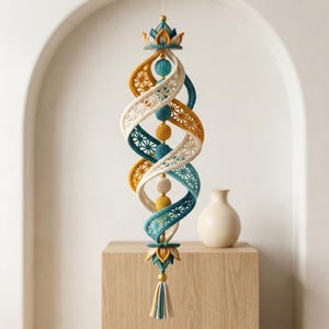 May include: A hanging spiral mobile featuring crocheted ribbons in teal, mustard yellow, and cream with intricate star-like patterns. The structure is accented by a central column of crocheted spheres and topped and bottomed with lotus-shaped elements.