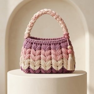 May include: A small, rounded handbag crafted from thick, braided yarn in a gradient of purple, pink, and cream tones. The bag features a single braided handle and a decorative side tassel with a small wooden bead accent, showcasing a textured, chunky knit pattern throughout the exterior.