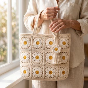 May include: Cream-colored crochet tote bag with daisy flower squares. The bag has a matching daisy flower charm with a tassel. The daisies have yellow centers and white petals. The bag is held by a person.