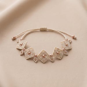 Pode incluir: Uma pulseira de macramé feita de cordão cor de creme, com cinco padrões tecidos em forma de diamante. Cada diamante contém uma conta central de metal em ouro rosa, com contas mais pequenas a acentuar as ligações e as extremidades do cordão.
