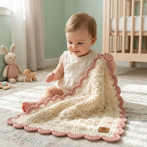 May include: A square crochet baby blanket featuring a cream-colored center with a textured star-like stitch pattern and a scalloped border in a dusty rose pink yarn. A small rectangular brown leather-like tag is attached to the bottom corner.