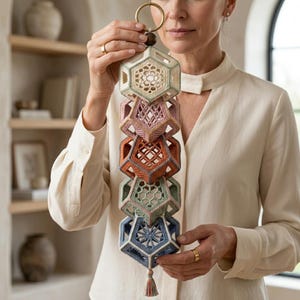 May include: A vertical hanging ornament composed of five stacked, crocheted geometric polyhedrons in shades of cream, dusty pink, terracotta, sage green, and slate blue. Each section features intricate openwork patterns and a small tassel at the base.