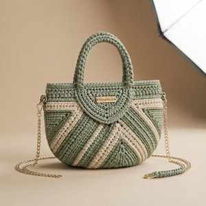 May include: A sage green and cream crochet handbag with a semi-circular shape, featuring a chevron pattern, two top handles, and a gold-tone metal chain shoulder strap. A small rectangular tag with the word Handmade is attached to the front.