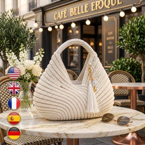 Puede incluir: Un bolso hobo de color crema hecho de cuerda gruesa enrollada con un asa redondeada. El bolso presenta un dije decorativo en tono dorado con una borla blanca larga unida al lateral. La textura está definida por crestas horizontales onduladas.