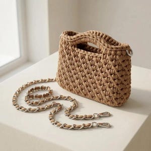 Pode incluir: Uma bolsa de mão em crochê bege com um padrão de malha espessa e texturizada com alças integradas. A bolsa inclui uma alça de ombro removível feita de elos de corrente de metal prateado entrelaçados com tiras de tecido bege correspondentes.