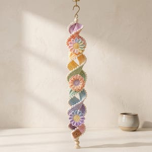 May include: A hanging spiral decoration made of crocheted yarn in pastel shades of pink, yellow, green, blue, and purple. The spiral features three integrated flower motifs and is edged with small white pom-poms, suspended from a gold metal hook.