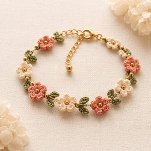 May include: A delicate bracelet featuring crocheted flowers in shades of pink and cream, interspersed with green leaves and gold beads. The bracelet has a gold-colored chain and clasp, creating a floral accessory.