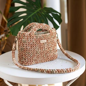 May include: A structured handbag crafted from thick, variegated yarn in shades of peach, cream, and sage green. The bag features two integrated top handles, a detachable braided shoulder strap with metal clasps, and a small rectangular label.