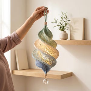 May include: A spiral-shaped hanging ornament crafted from crocheted yarn in gradient shades of sage green, cream, and light blue. The structure features an openwork pattern, a braided loop at the top, and a clear faceted crystal bead at the bottom.