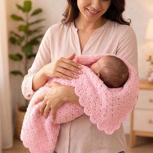 May include: A baby wrapped in a soft, light pink crocheted blanket with a scalloped edge. The blanket is held by a person wearing a light pink shirt.