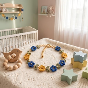 May include: A delicate bracelet featuring alternating blue and yellow crocheted flowers with small green leaves and gold-toned beads. The piece includes a gold-colored metal chain with a lobster clasp closure, arranged in a circular pattern on a textured fabric.