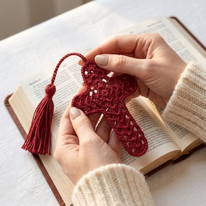 Può includere: Segnalibro a forma di croce rossa all'uncinetto fatto a mano con nappa. Il segnalibro è tenuto aperto in un libro con la parola "Matthew" visibile. La croce ha un design traforato.