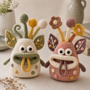 Peut inclure: Deux créatures en crochet avec de grands yeux et de larges bouches. La figurine de gauche est crème avec des accents verts et jaunes, tandis que celle de droite est rose poudré avec des détails marron et jaunes. Elles ont des tiges florales.