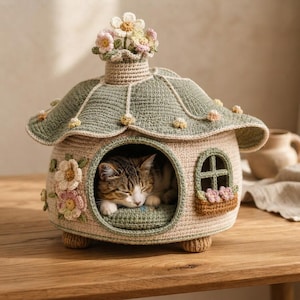 May include: A beige and sage green crocheted cat house shaped like a cottage with a wide roof, a central arched entrance, and a small window. The roof is topped with crocheted flowers, and the exterior features floral accents and small feet.