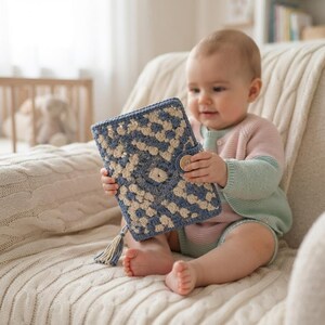 May include: A rectangular crochet book cover featuring a blue and cream geometric pattern. The cover includes a small wooden button closure on the side and a decorative cream-colored tassel hanging from the bottom corner of the textured fabric.