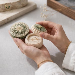 May include: A round, crocheted ring box in cream and sage green with a floral pattern on the lid. The open box reveals a beige velvet interior holding a thin gold ring with a small clear stone. The lid is held separately beside the base.