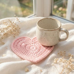 May include: A light pink heart-shaped crochet coaster made of yarn with a textured stitch pattern. A speckled cream-colored ceramic mug with a handle sits on top of the coaster. The items are placed on a white fabric surface with dried flowers.