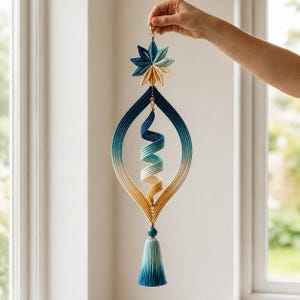 May include: A hanging decorative ornament featuring a star-shaped top, a central spiral element, and a bottom tassel. The piece is crafted from thread in a gradient color scheme transitioning from dark blue to teal, light blue, and warm yellow.