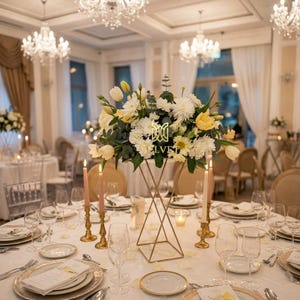 Puede incluir: Una mesa de comedor formal con un soporte floral geométrico dorado que contiene flores blancas y amarillas. La mesa está puesta con platos con borde dorado, velas y cristalería. La habitación tiene candelabros y una decoración elegante.