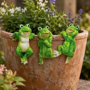 May include: Three green frog ornaments with white bellies and black eyes cling to the rim of a terracotta plant pot. The frogs appear to be climbing or hanging on the pot, adding a whimsical touch to the garden, or patio.