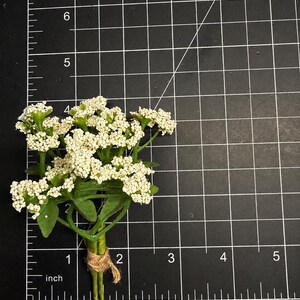May include: A small artificial flower bouquet featuring clusters of tiny white blossoms and green leaves, tied together with a piece of natural twine. The bouquet measures approximately 2.5 inches in height against a black grid mat.