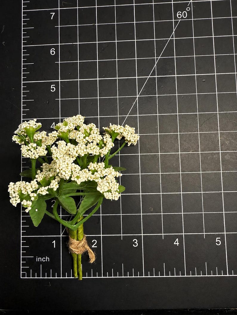 May include: A small artificial flower bouquet featuring clusters of tiny white blossoms and green leaves, tied together with a piece of natural twine. The bouquet measures approximately 2.5 inches in height against a black grid mat.