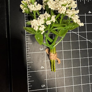 May include: A small artificial flower bouquet with white clusters and green leaves, tied with brown twine. The bouquet is positioned on a black cutting mat with white grid lines, showing a length of approximately 3.5 inches from top to bottom.