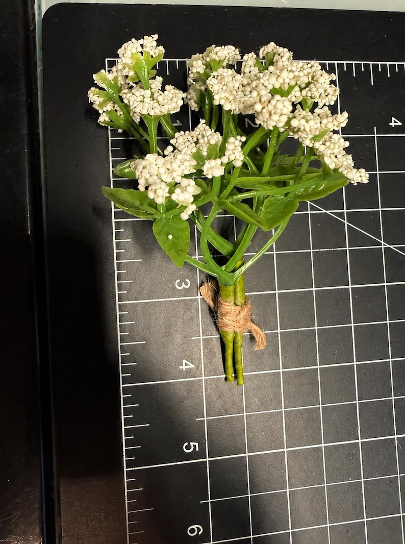 May include: A small artificial flower bouquet with white clusters and green leaves, tied with brown twine. The bouquet is positioned on a black cutting mat with white grid lines, showing a length of approximately 3.5 inches from top to bottom.