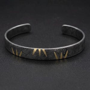 Puede incluir: Una pulsera rígida de metal gris oscuro con un patrón topográfico complejo y ondulado en toda su superficie. La pulsera está decorada con varias incrustaciones triangulares pequeñas, afiladas y de color dorado dispuestas en grupos.