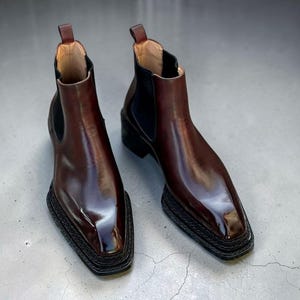 May include: A pair of dark brown leather Chelsea boots with a polished finish, featuring a square toe, black elastic side panels, a pull tab at the heel, and a thick, textured black rubber lug sole with a prominent welt edge.