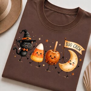 May include: A brown t-shirt featuring a graphic of a black cat wearing a witch hat, a piece of candy corn, a caramel apple, and a crescent moon holding a flag that reads BOO CREW, all decorated with small stars on the fabric surface.