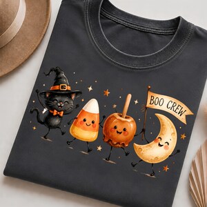 May include: A dark gray t-shirt featuring a graphic of a black cat wearing a witch hat, a piece of candy corn, a caramel apple, and a crescent moon, all with smiling faces. The moon holds a banner with the text BOO CREW, surrounded by stars.
