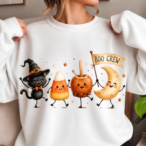 May include: A white sweatshirt featuring a graphic of a black cat in a witch hat, a piece of candy corn, a caramel apple, and a crescent moon holding a flag that reads BOO CREW. The characters have smiling faces and are surrounded by small stars.